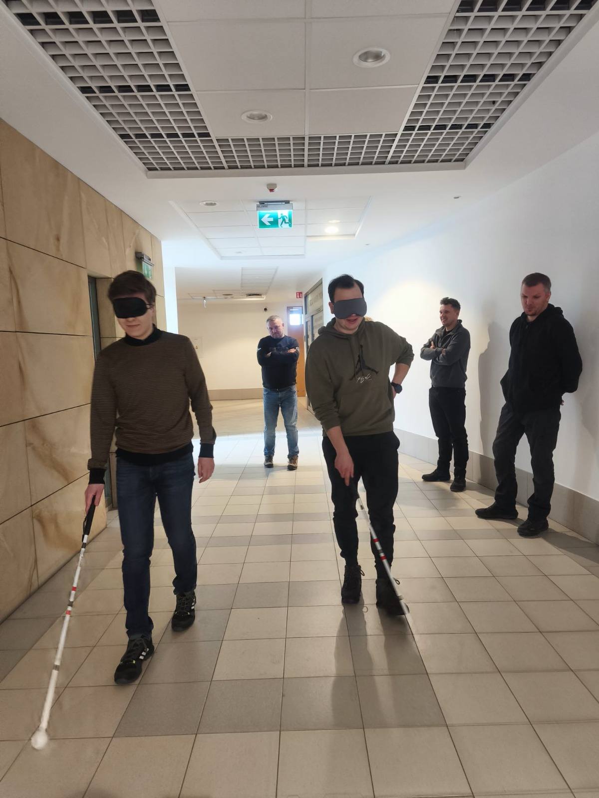 Osoby z zasłoniętymi oczami i białymi laskami spacerujący po korytarzu.